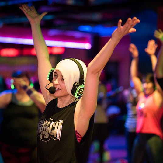 Quiet Events Silent Disco headphones for fitness and meditation.  A fitness instructor providing a class to her students with her hands up and wearing headphones.