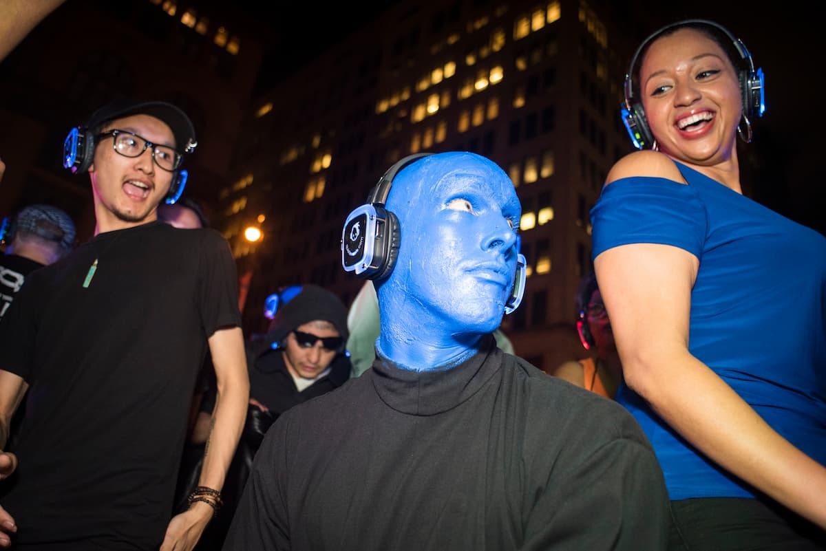 Quiet Events Silent Disco headphones for public spaces a blue man kneel down in between two people wearing headphones 