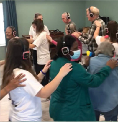 Quiet Events Silent Disco headphones for senior care.  A group of older people walking in a circle holding there backs with hands and wearing headphones.