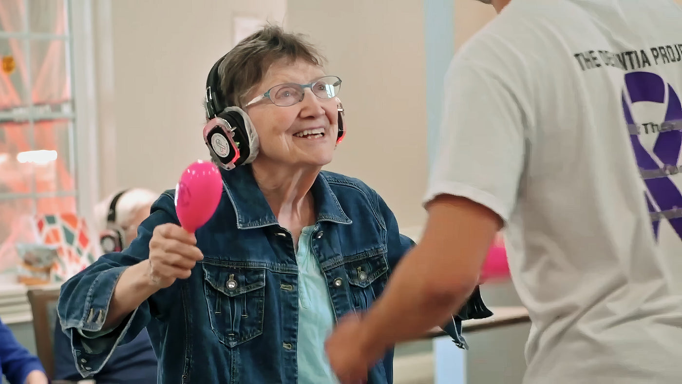 Quiet Events Silent Disco headphones for senior care.  A older woman having fun being entertained holding maracas and wearing headphones.