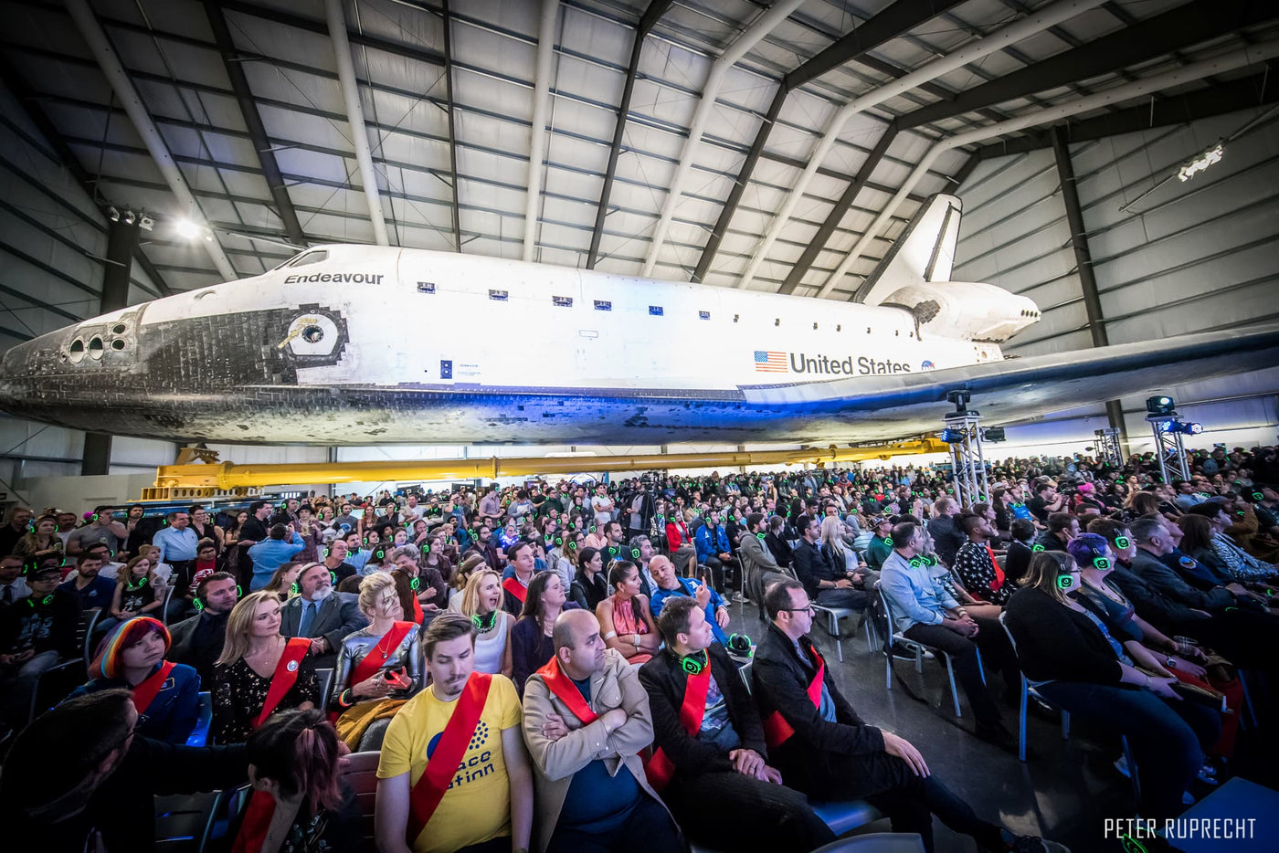 Silent Disco headphones for other events.  A group of people gather and listening carefully during a presentation with a NASA space shuttle suspend in the air wearing headphones.