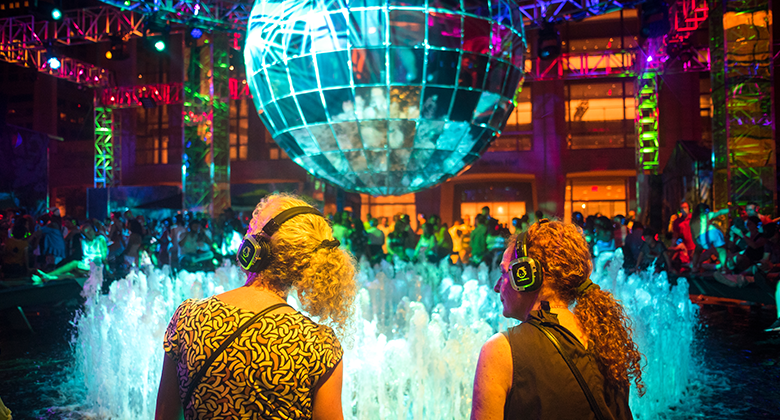 Two people looking up at a disco ball wearing glowing headphones at a silent disco